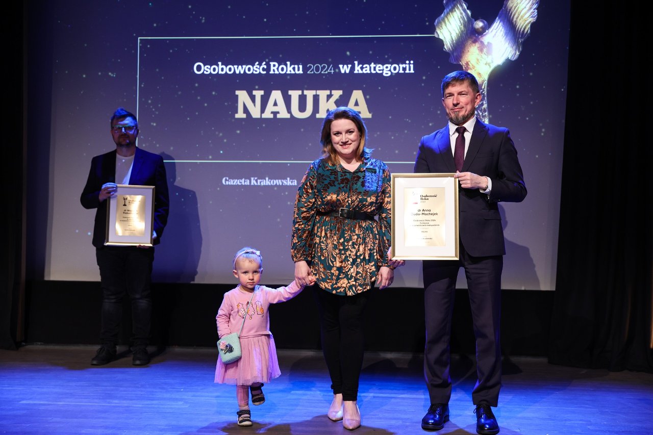 Trzy osoby stoją na scenie podczas gali „Osobowość Roku 2024”. W centrum stoi uśmiechnięta kobieta w kwiecistej sukience, trzymając za rękę małą dziewczynkę w różowej sukience. Po prawej stronie mężczyzna w garniturze trzyma oprawiony dyplom. Na ekranie w tle widnieje napis „Osobowość Roku 2024 w kategorii NAUKA”.