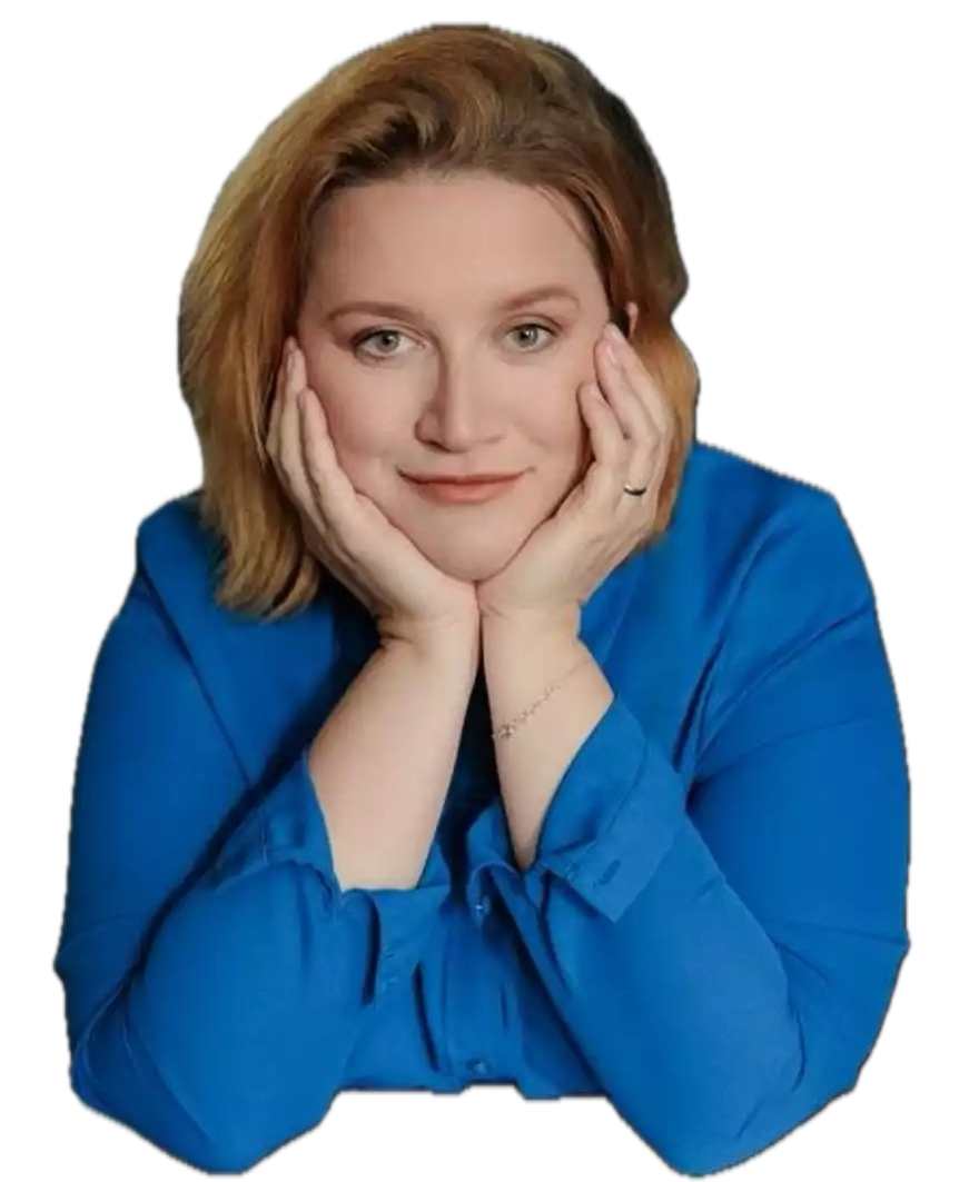 Portret uśmiechniętej kobiety o rudych włosach, ubranej w niebieską koszulę. Kobieta opiera podbródek na złączonych dłoniach, patrząc bezpośrednio w stronę obiektywu na białym tle.